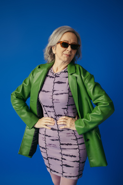 senior woman in green leather jacket and dress standing with hands on hips isolated on blue - Photo, Image