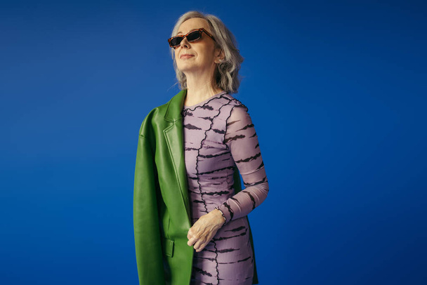 senior woman in sunglasses and dress standing with green leather jacket isolated on blue - Photo, image