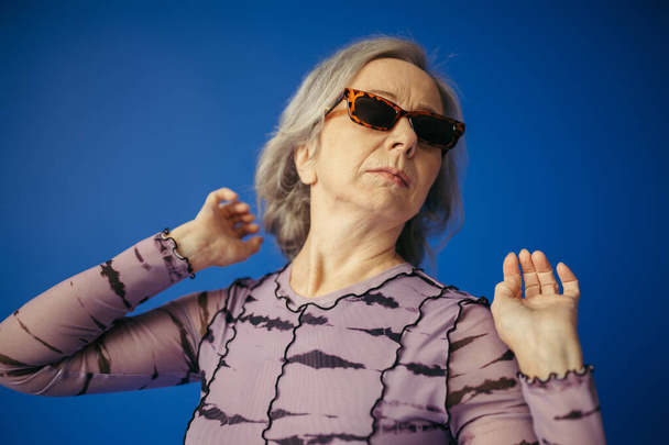 Seniorin mit stylischer Sonnenbrille posiert isoliert auf blauem Grund - Foto, Bild