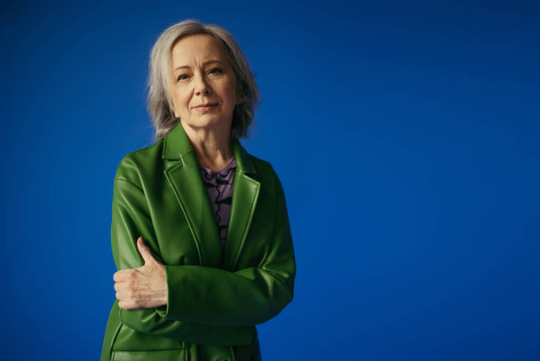 positive grey haired woman in green leather jacket standing with crossed arms isolated on blue - Photo, image