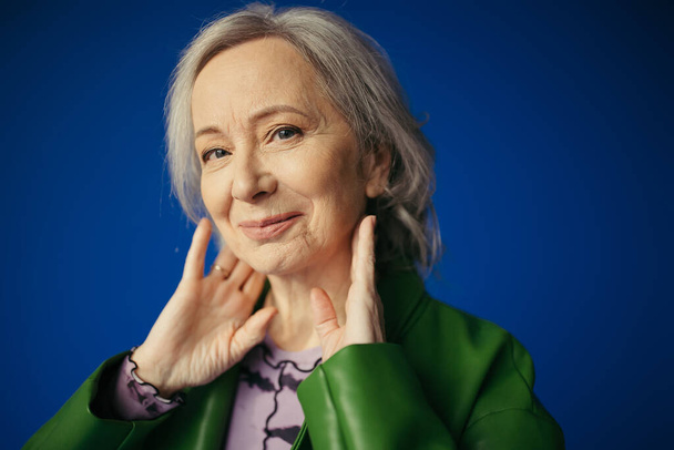 muotokuva positiivinen ja tyylikäs vanhempi nainen poseeraa kädet lähellä kaulaa ja hymyillen kameran eristetty sininen - Valokuva, kuva