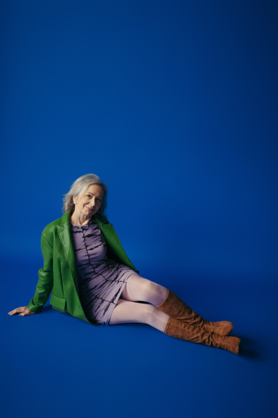 full length of happy senior model in stylish casual attire sitting on blue background - Fotó, kép