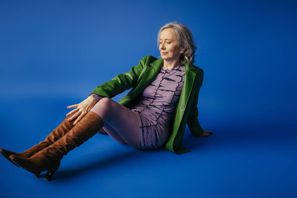full length of trendy senior woman in suede boots and green leather jacket over lilac dress sitting on blue background - Fotó, kép