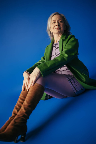 lachend senior model in groen leren jasje en bruine suède laarzen zittend op blauwe achtergrond - Foto, afbeelding