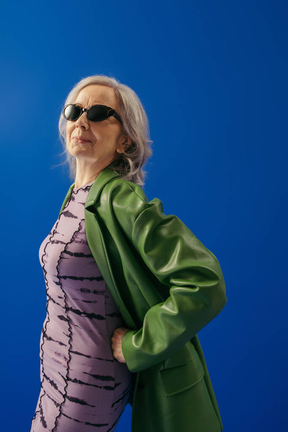 grey haired woman in trendy sunglasses and green leather jacket over purple dress posing with hand on hip isolated on blue - Foto, imagen