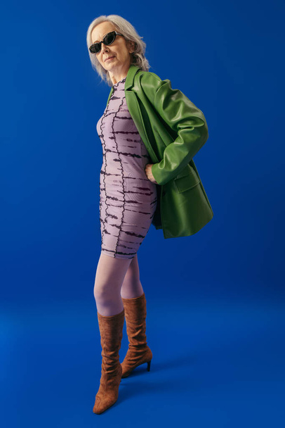 elegant senior lady in suede boots and green leather jacket over purple dress standing with hand on hip on blue background - 写真・画像