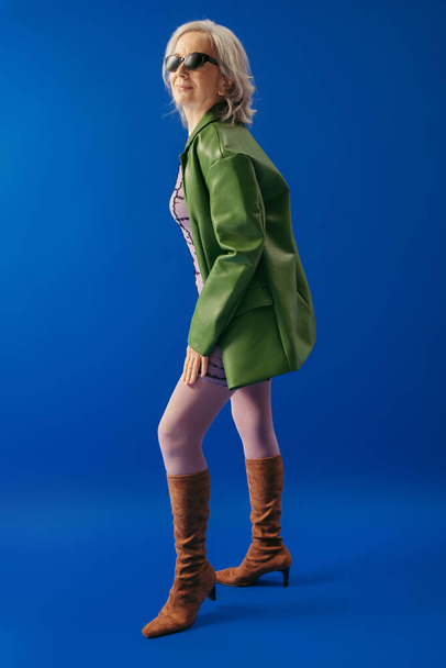 full length of positive senior woman in sunglasses and green leather jacket with brown suede boots on blue background - Zdjęcie, obraz