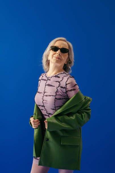 donna dai capelli grigi in occhiali da sole alla moda e abito lilla con giacca di pelle verde isolato su blu - Foto, immagini