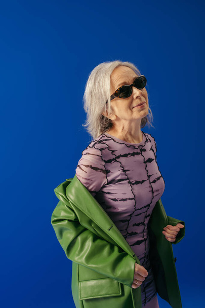 старшая женщина в солнцезащитных очках и зеленой кожаной куртке поверх фиолетового платья улыбается изолированы на синий - Фото, изображение