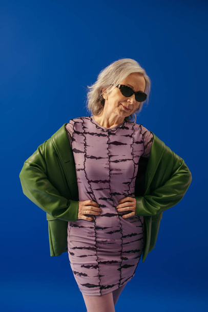 smiling senior model in sunglasses and green leather jacket over purple dress posing with hands on waist isolated on blue - Photo, Image