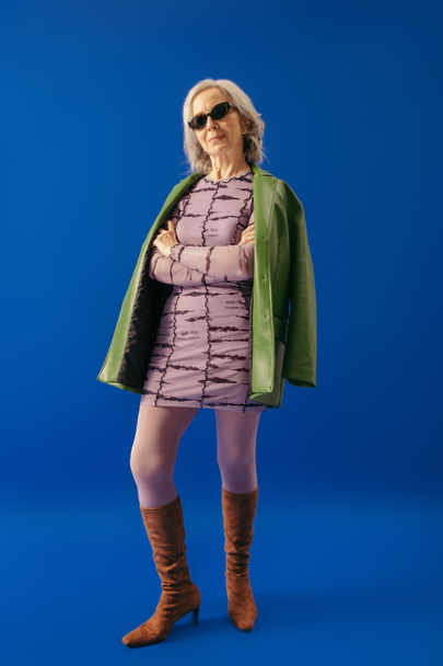 elegant grey haired woman in suede boots and green leather jacket over purple dress standing with crossed arms on blue  - Photo, Image