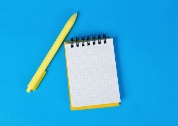 Κίτρινο σημειωματάριο με χαλαρό χαρτί και ένα στυλό σε μπλε φόντο. Κενή σελίδα σε ένα κελί για κείμενο. Άποψη από ψηλά. - Φωτογραφία, εικόνα