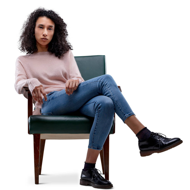 Mladá kudrnatá žena seděla na židli se zkříženýma nohama a s výrazem vážné sebedůvěry. Vyzývá tě. Izolováno na průhledné nebo bílé. - Fotografie, Obrázek