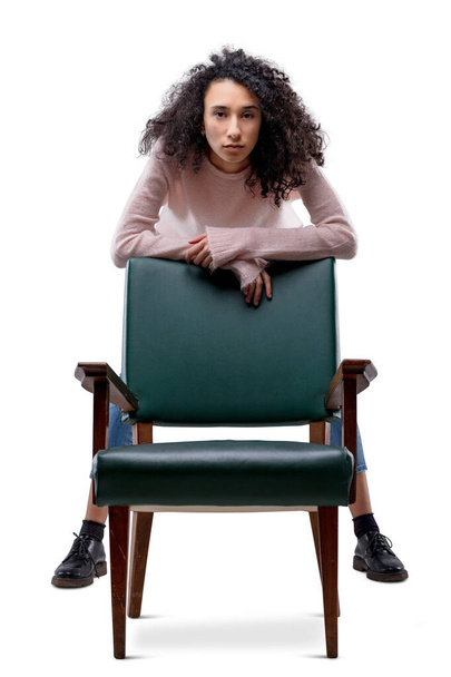 Young curly-haired woman rests her arms on a chair that stands in front of her like a shield and lets her keep her long legs straight. She wears a pink sweater and jeans. Isolated on transparent or wh - Foto, Bild