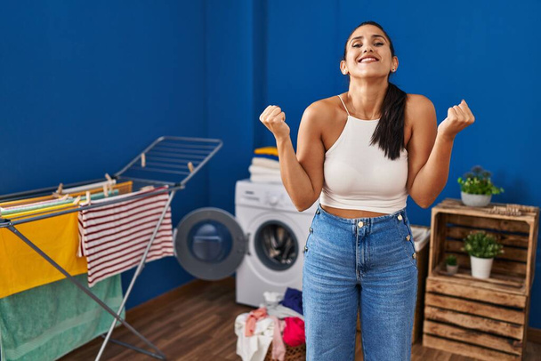 Joven mujer hispana en la lavandería gritando orgullosa, celebrando la victoria y el éxito muy emocionada con los brazos levantados  - Foto, imagen