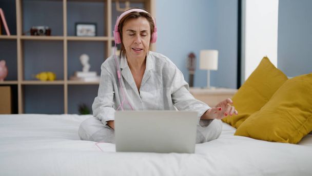 Middle age hispanic woman listening to music sitting on bed at bedroom - Фото, изображение