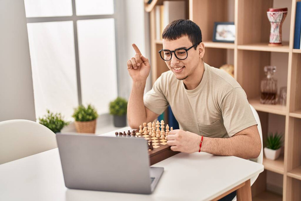 młody arabski człowiek gra w szachy online uśmiechnięty szczęśliwy wskazując ręką i palcem na bok  - Zdjęcie, obraz