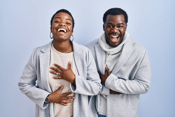 Nuori afrikkalainen amerikkalainen pari seisoo sinisellä taustalla yhdessä hymyillen ja nauraen kovaa ääneen, koska hauska hullu vitsi kädet vartalolla.  - Valokuva, kuva