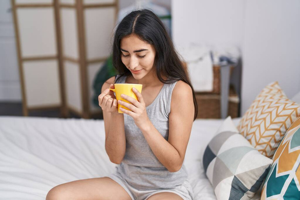 Young hispanic girl drinking cup of coffee sitting on bed at bedroom - Foto, Imagem