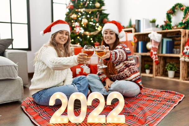 Woman couple toasting with champagne for new year 2022 sitting by christmas tree at home - Photo, Image