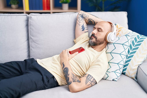 Young bald man listening to music sleeping on sofa at home - Foto, imagen