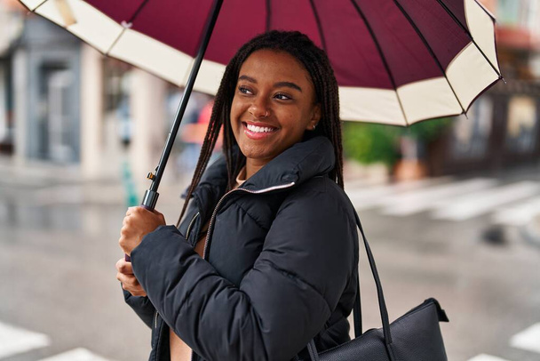 African american woman smiling confident using umbrella at street - Photo, Image