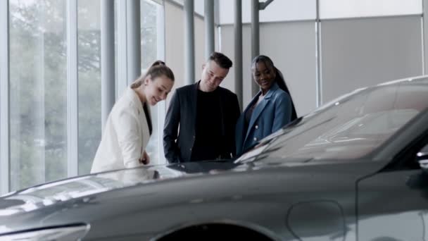 Los clientes que inspeccionan el coche nuevo en un concesionario moderno. Un vendedor atractivo habla de un coche eléctrico y disfruta del hermoso coche - Imágenes, Vídeo