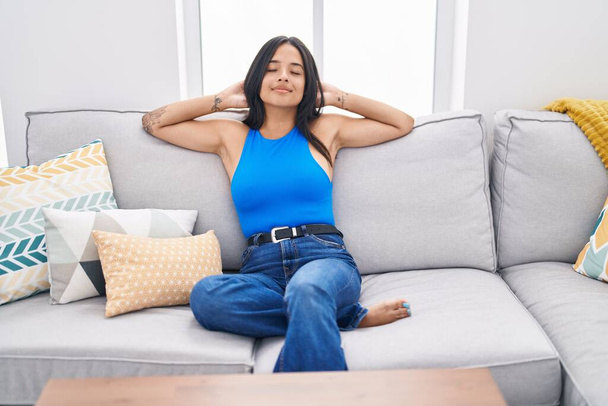 Young hispanic woman relaxed with hands on head sitting on sofa at home - Foto, imagen