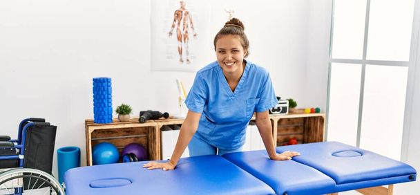 Jovem bela mulher hispânica fisioterapeuta sorrindo confiante em pé na clínica de reabilitação - Foto, Imagem
