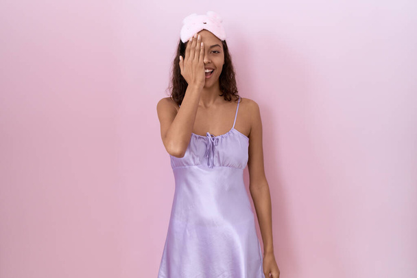 Young hispanic woman wearing sleep mask and nightgown covering one eye with hand, confident smile on face and surprise emotion.  - Foto, immagini