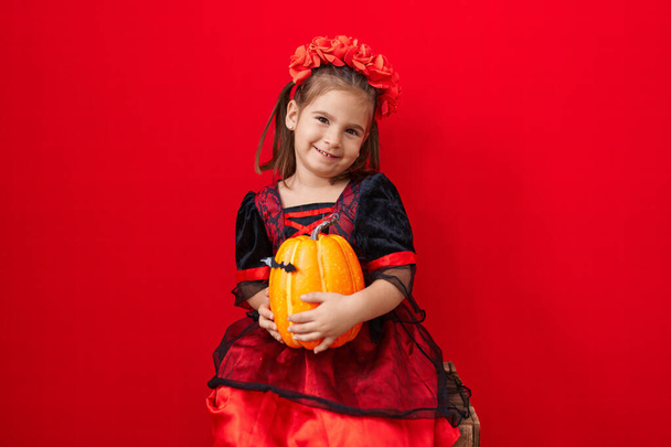 Adorable hispanic girl wearing halloween costume holding pumpkin over isolated red background - 写真・画像