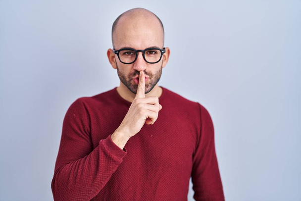 Jeune homme chauve avec la barbe debout sur fond blanc portant des lunettes demandant à être calme avec le doigt sur les lèvres. silence et concept secret.  - Photo, image