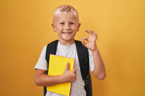 Mały chłopiec kaukaski nosząc plecak studencki i trzymając książkę robi ok znak palcami, uśmiechając przyjazny gest doskonały symbol  - Zdjęcie, obraz