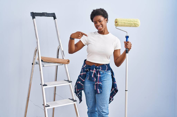 Mujer afroamericana sosteniendo rodillo pintor mirando confiado con sonrisa en la cara, señalándose con los dedos orgullosos y felices.  - Foto, imagen