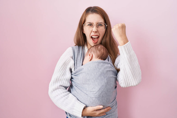 Young caucasian woman holding and carrying baby on a sling annoyed and frustrated shouting with anger, yelling crazy with anger and hand raised  - Φωτογραφία, εικόνα