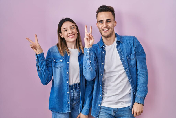 Молодая латиноамериканская пара, стоящая на розовом фоне и улыбающаяся со счастливым лицом, подмигивая в камеру, делая знак победы пальцами. Номер два.  - Фото, изображение