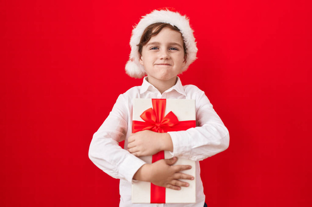 Маленький білий хлопчик носить різдвяний капелюх і тримає подарунки набиваючи щоки смішним обличчям. рот, надутий повітрям, ловить повітря
.  - Фото, зображення