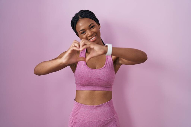 Donna afroamericana con trecce che indossano abbigliamento sportivo su sfondo rosa sorridente in amore facendo forma di simbolo del cuore con le mani. concetto romantico.  - Foto, immagini