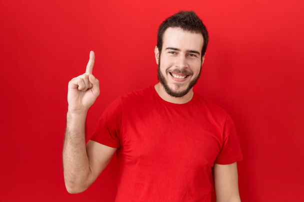 Молодой латиноамериканец в непринужденной красной футболке показывает и показывает пальцем номер один, улыбаясь уверенно и счастливо.  - Фото, изображение