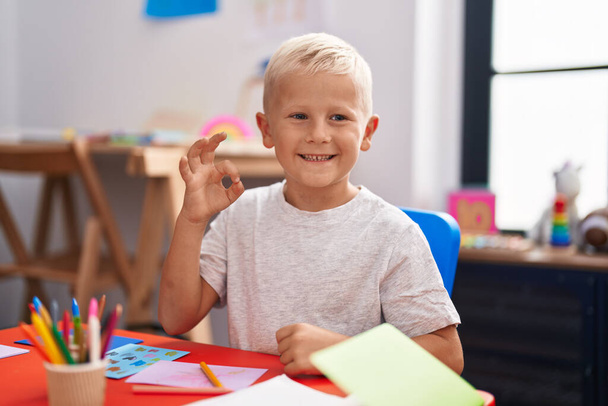 Kleiner kaukasischer Junge, der in der Schule malt und mit den Fingern ein Zeichen macht, lächelt freundlich und gestikuliert ein ausgezeichnetes Symbol  - Foto, Bild