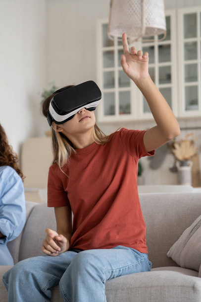 Curious teen girl child sitting on sofa using VR headset playing games at home while her mother working beside, kid touching objects in immersive virtual reality, children exploring augmented reality - Photo, image