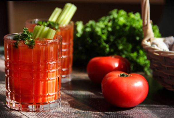 tomato juice close up with fresh tomatoes and basil on a dark background. two glasses of bloody mary cocktail with fresh celery and parsley on a wooden table in natural lightning, rustic style. Fresh vegetable juice with tomatoes - Fotografie, Obrázek