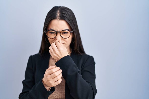 Young brunette woman standing over blue background smelling something stinky and disgusting, intolerable smell, holding breath with fingers on nose. bad smell  - Фото, изображение