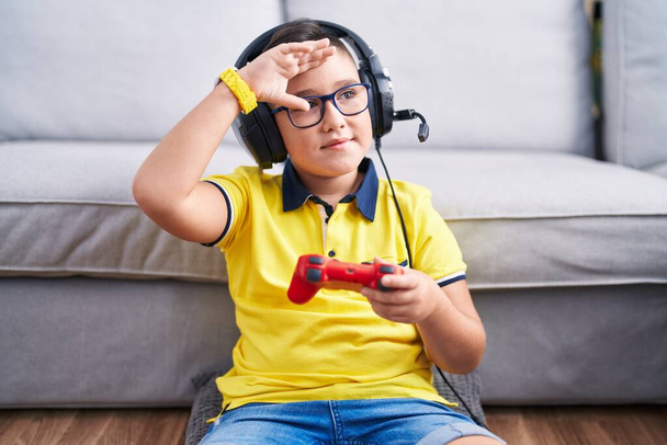 Young hispanic kid playing video game holding controller wearing headphones very happy and smiling looking far away with hand over head. searching concept.  - Photo, image