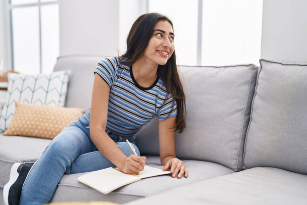 Молода іспаномовна дівчина пише на блокноті, сидячи на дивані вдома
 - Фото, зображення