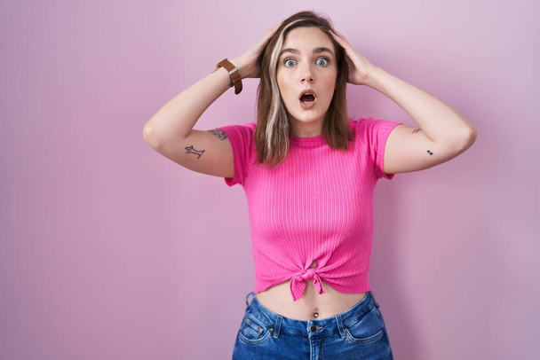 Blonde caucasian woman standing over pink background crazy and scared with hands on head, afraid and surprised of shock with open mouth  - Fotografie, Obrázek