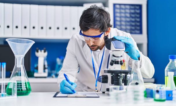 Jonge Spaanse man wetenschapper met behulp van microscoop schrijven op klembord in het laboratorium - Foto, afbeelding