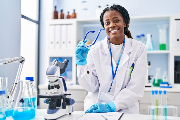 Afrikaans amerikaanse vrouw wetenschapper glimlachen zelfverzekerd holding veiligheidsbril in laboratorium - Foto, afbeelding