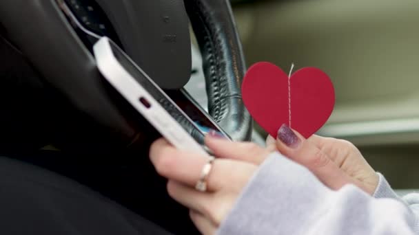 молодая женщина, сидящая в кресле водителя современный автомобиль интерьер набрав сообщение с помощью смартфона держа бумажное сердце на другой hand.long светлые волосы готовы к романтической поездки в одиночку, руль - Кадры, видео