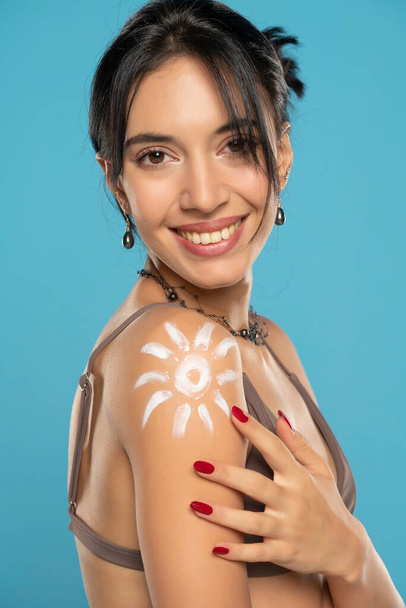 Happy Woman In Bikini With Suntan Lotion Shaped Sun On Her Shoulder, On A Blue Studio Background.  - 写真・画像